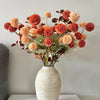 A bouquet of artificial chrysanthemum pompon flowers in shades of orange and rust arranged in a light beige vase.