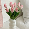A bouquet of six pink tulips in a white vase displayed on a white surface.