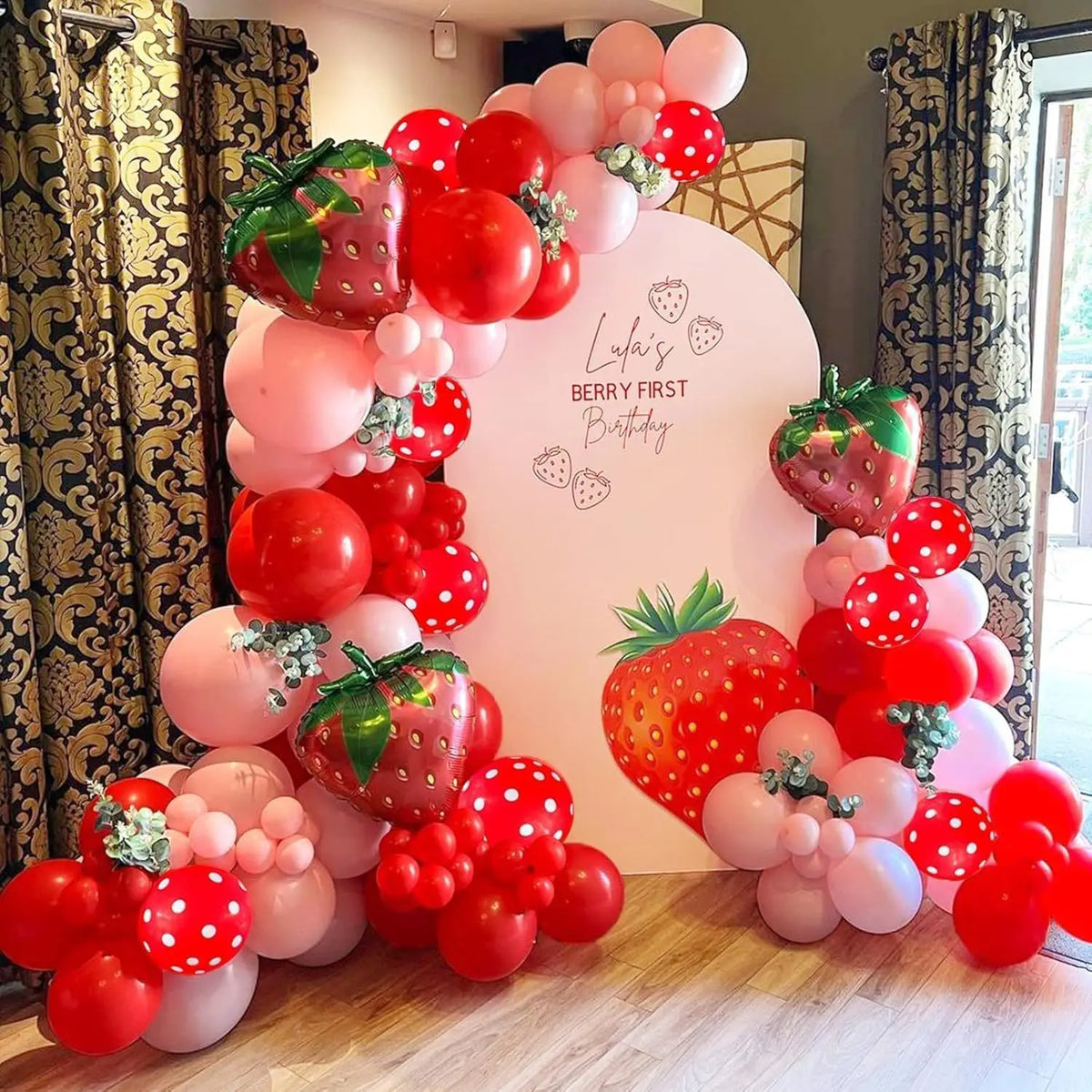 A pink and red balloon arch display featuring strawberries and the text 'Lufai's Berry First Birthday'.