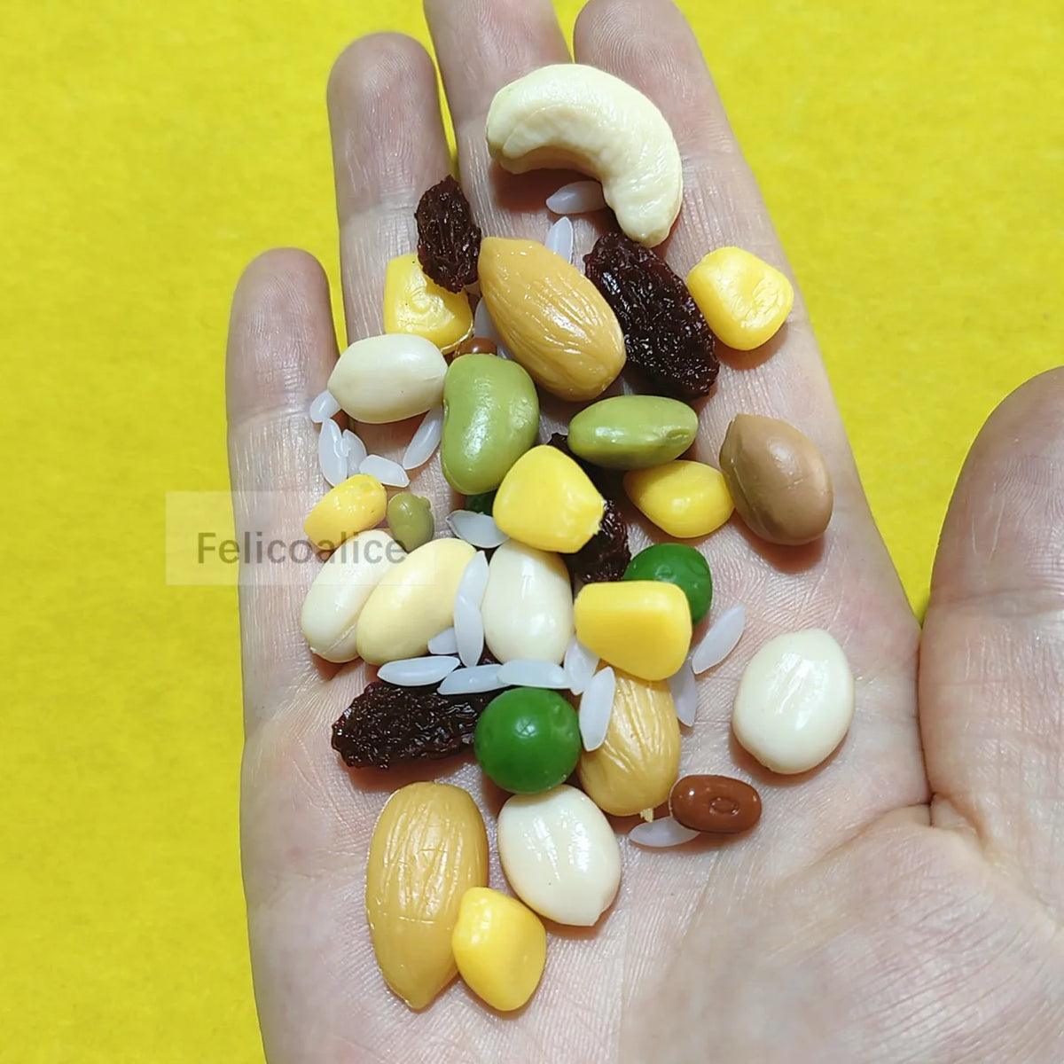 A hand holds various realistic resin fake corn miniatures in yellow, green, and beige colors on a yellow background.