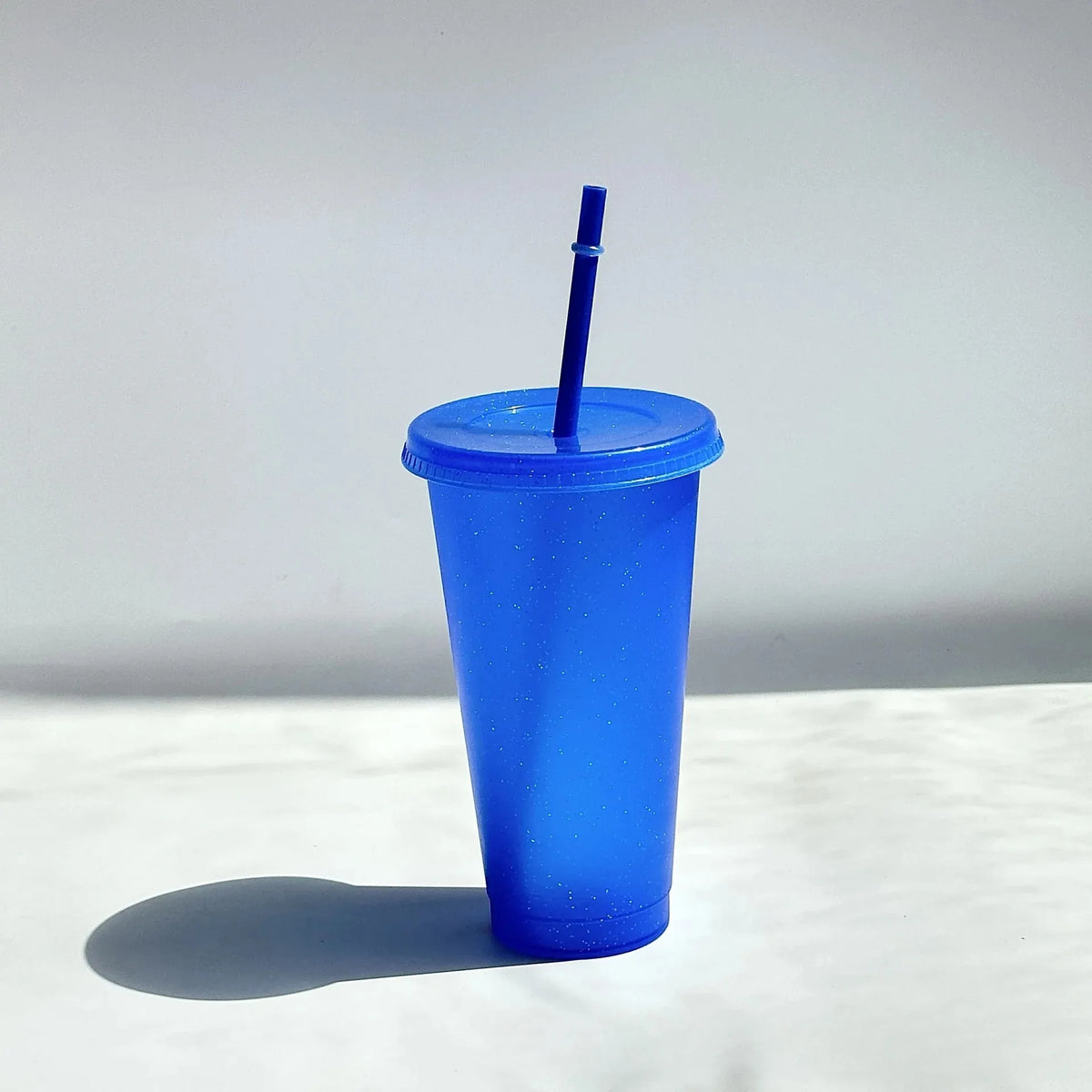 A bright blue glitter party cup with a straw is shown against a white background.
