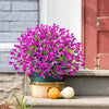 A vibrant bouquet of purple artificial calla lilies in a green pot sits on stone steps next to a pink wooden door.