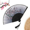 A black folding fan with gray ribs decorated with pink floral patterns and a bamboo texture visible in the background.