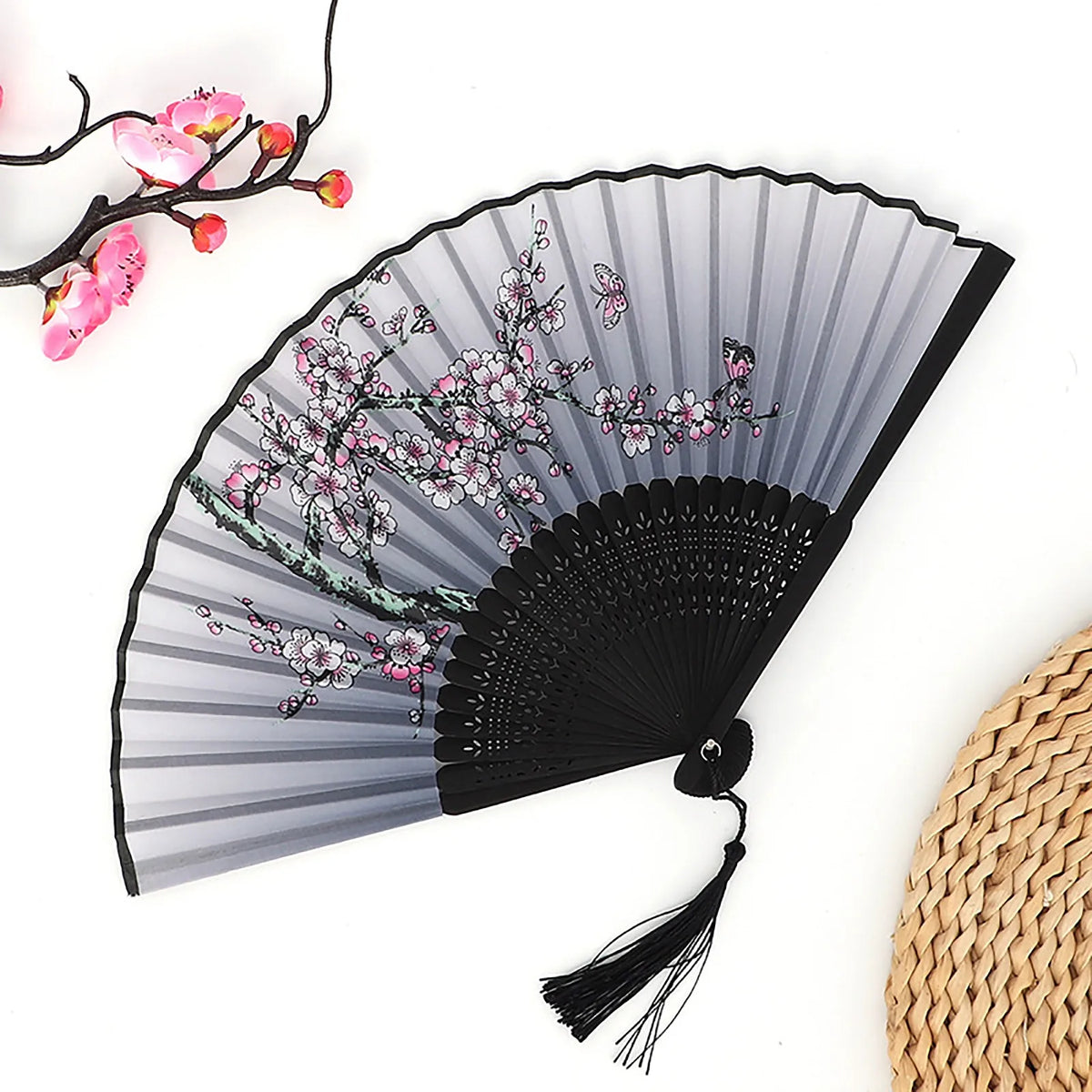 A black folding fan with gray ribs decorated with pink floral patterns and a bamboo texture visible in the background.