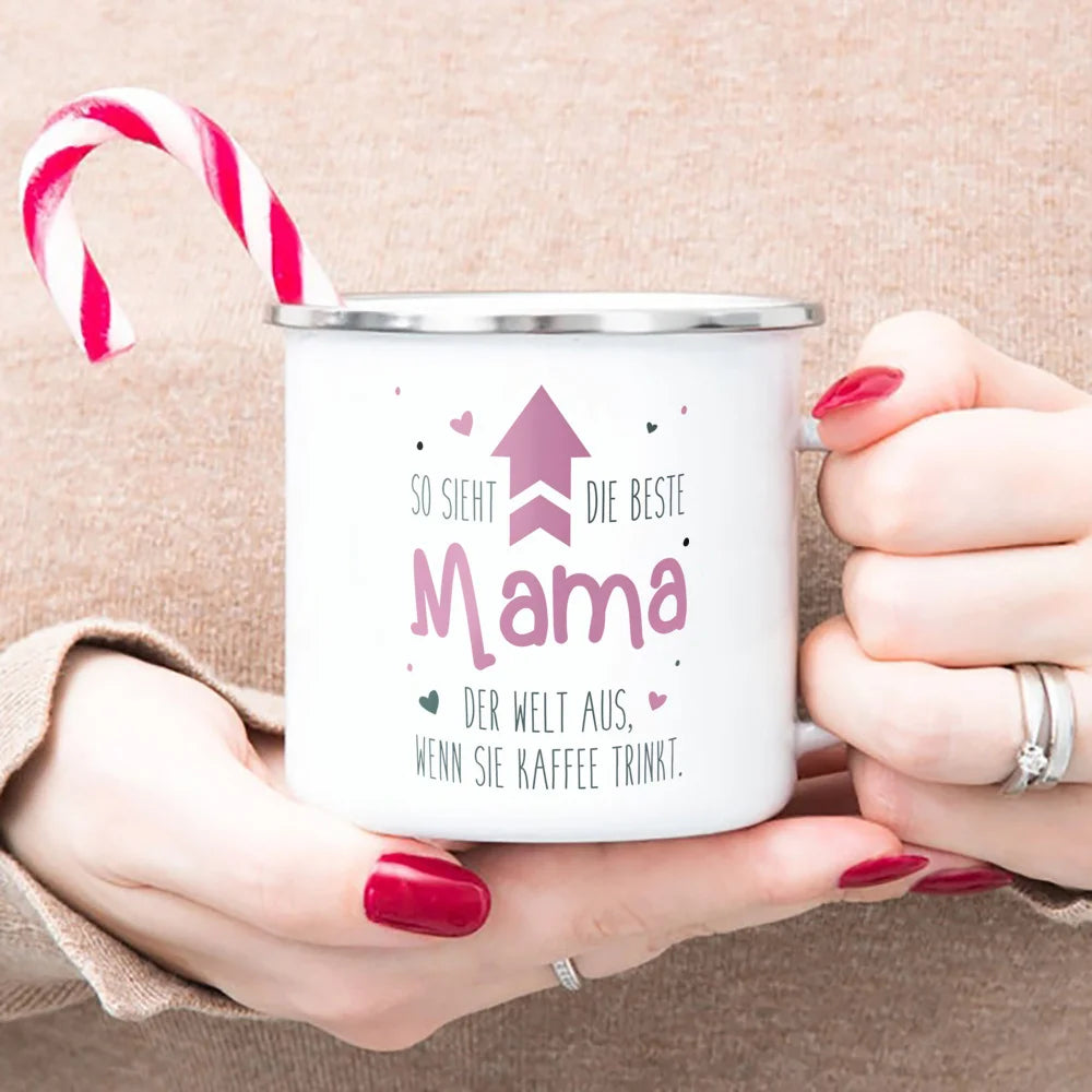 A hand holds a white enamel mug with pink text and a red and white striped candy cane resting on the rim.