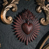 A dark wall plaque featuring a reddish-brown sacred heart design surrounded by radiating lines and ornate gold scrollwork.