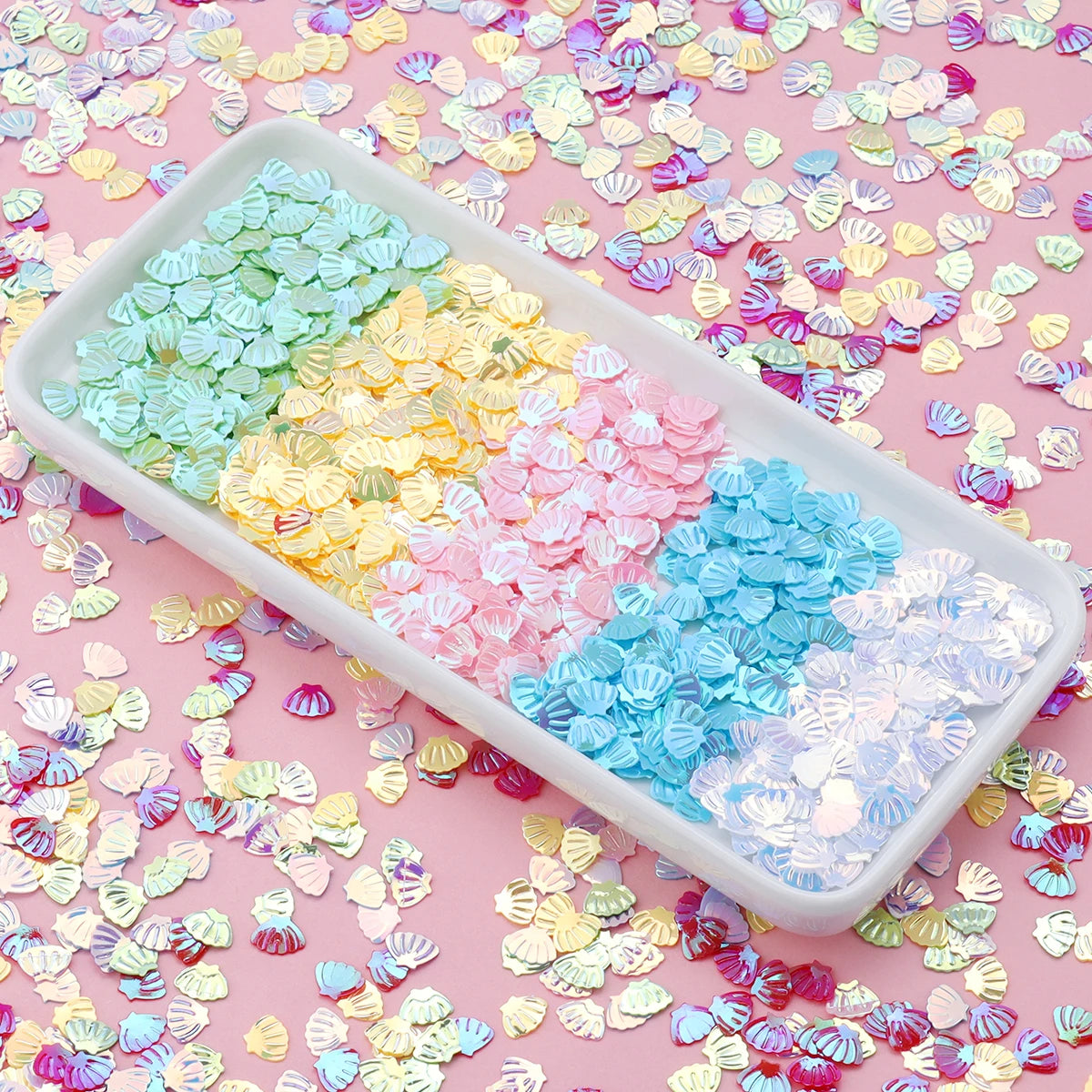A white dish filled with pink, blue, and yellow sea shell PVC sequins scattered on a pink background.