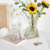A clear glass vase shaped like a money bag holds a bright yellow sunflower with green leaves.