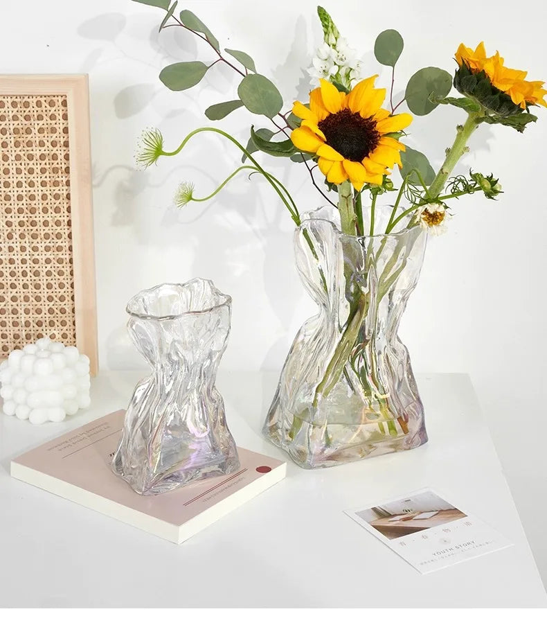A clear glass vase shaped like a money bag holds a bright yellow sunflower with green leaves.