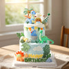 Pastel de cumpleaños de varios pisos decorado con figuras de dinosaurios y hojas tropicales sobre fondo blanco.
