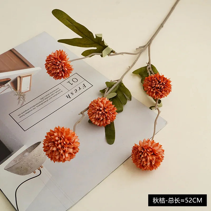 A sprig of artificial orange chrysanthemum pompon flowers with green leaves displayed on a white card.