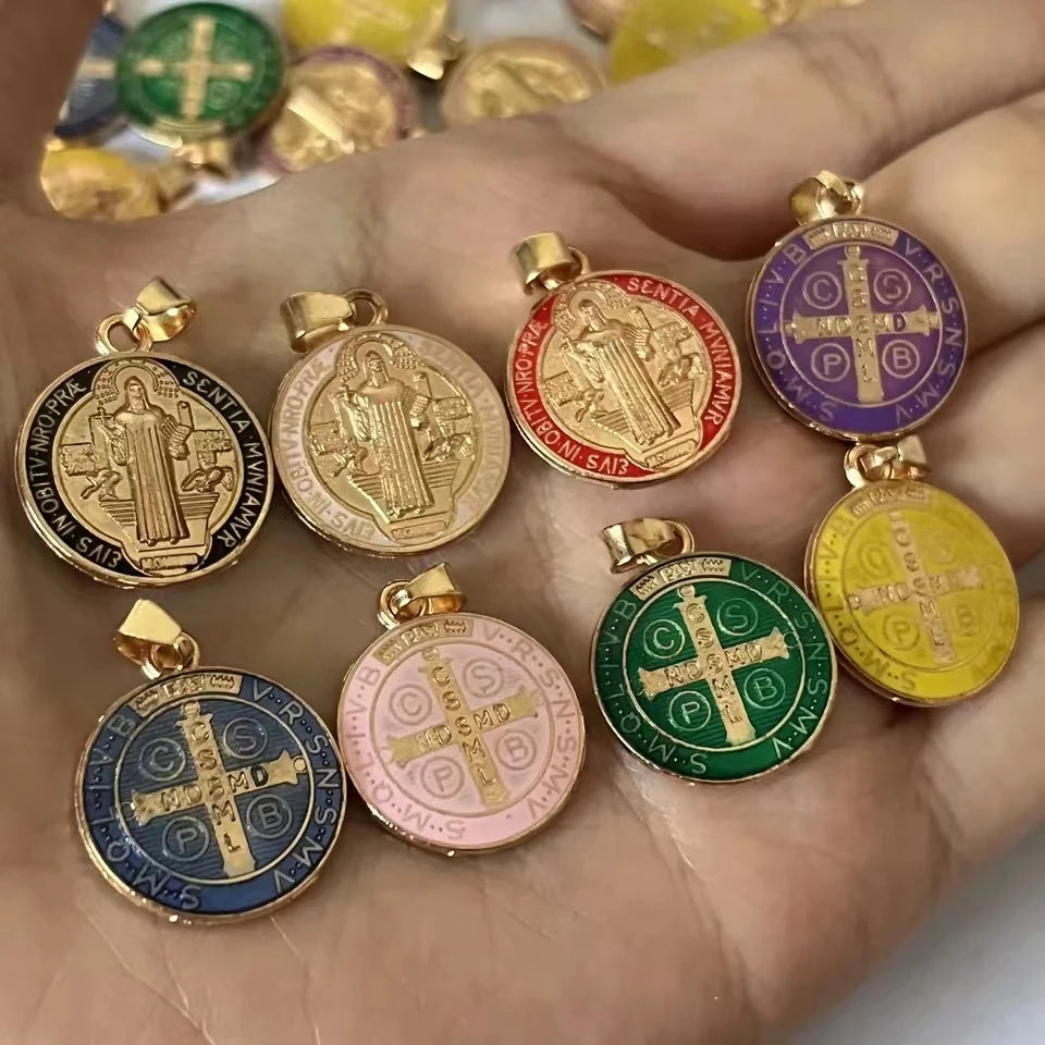 A hand holds a set of twelve double-sided St. Benedict medals in various colors and gold-toned metal.