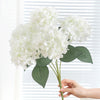 A hand holds a cluster of white artificial hydrangea silk flowers against a white window blind.