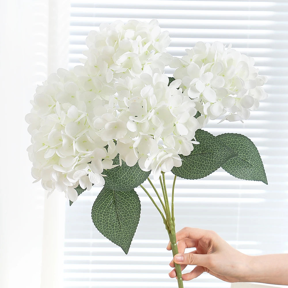 A hand holds a cluster of white artificial hydrangea silk flowers against a white window blind.