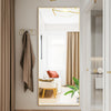 A modern arched full-length mirror with a gold frame is shown in a bright interior with neutral tones and sheer curtains.