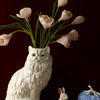 A white ceramic cat figurine with a vase of pink tulips and green leaves on a wooden surface.