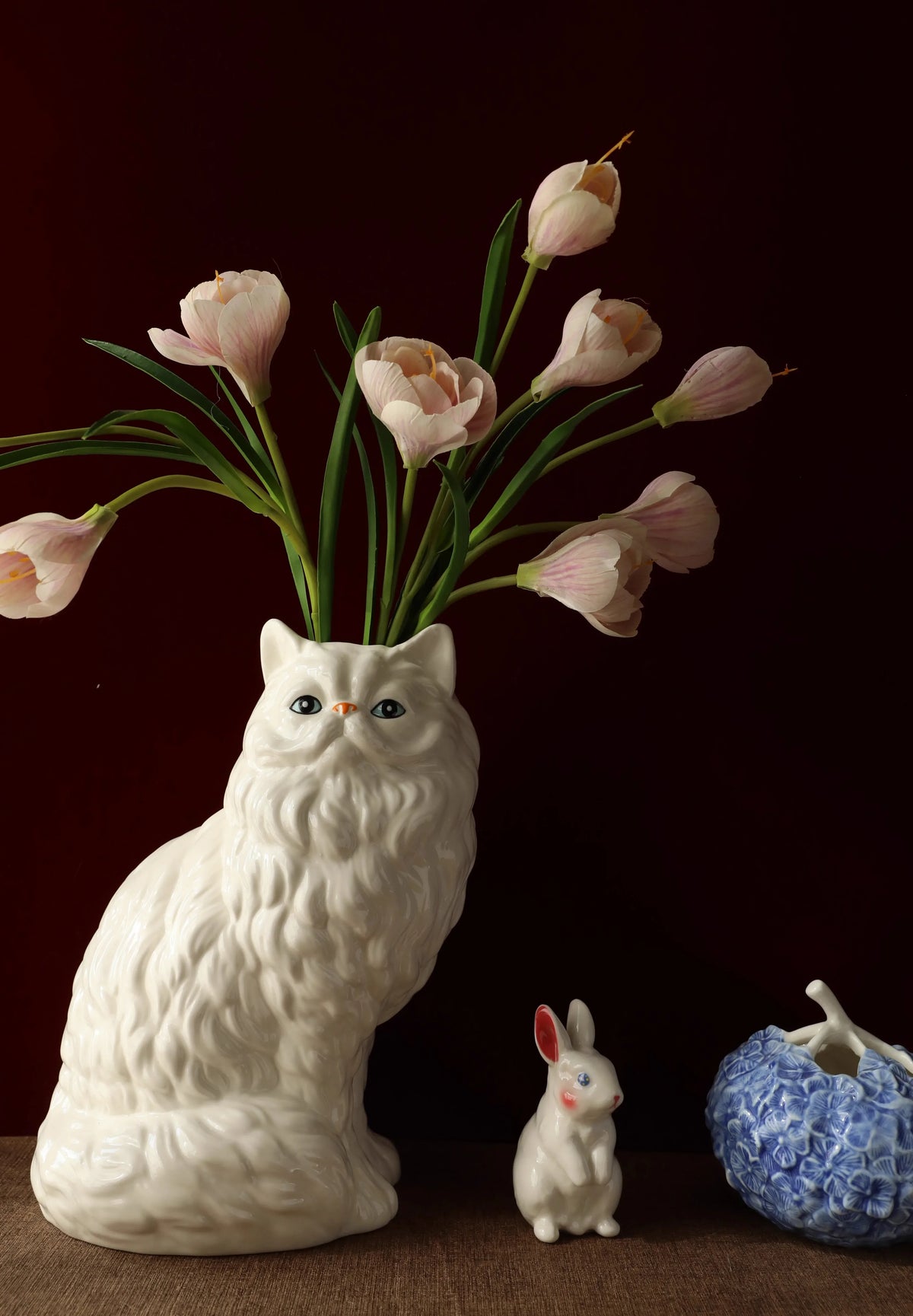 A white ceramic cat figurine with a vase of pink tulips and green leaves on a wooden surface.