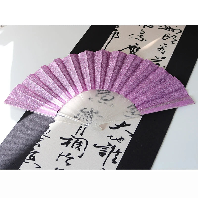 A purple glitter folding fan with transparent ribs is displayed against a black background and traditional Japanese writing.