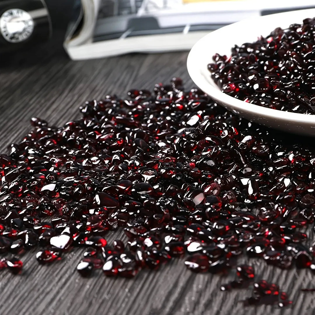 A pile of deep burgundy natural garnet crystal chips scattered on a dark surface with a small silver dish nearby.