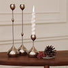 A set of three metallic waterdrop design candle holders with white candles and a pinecone on a wooden surface.