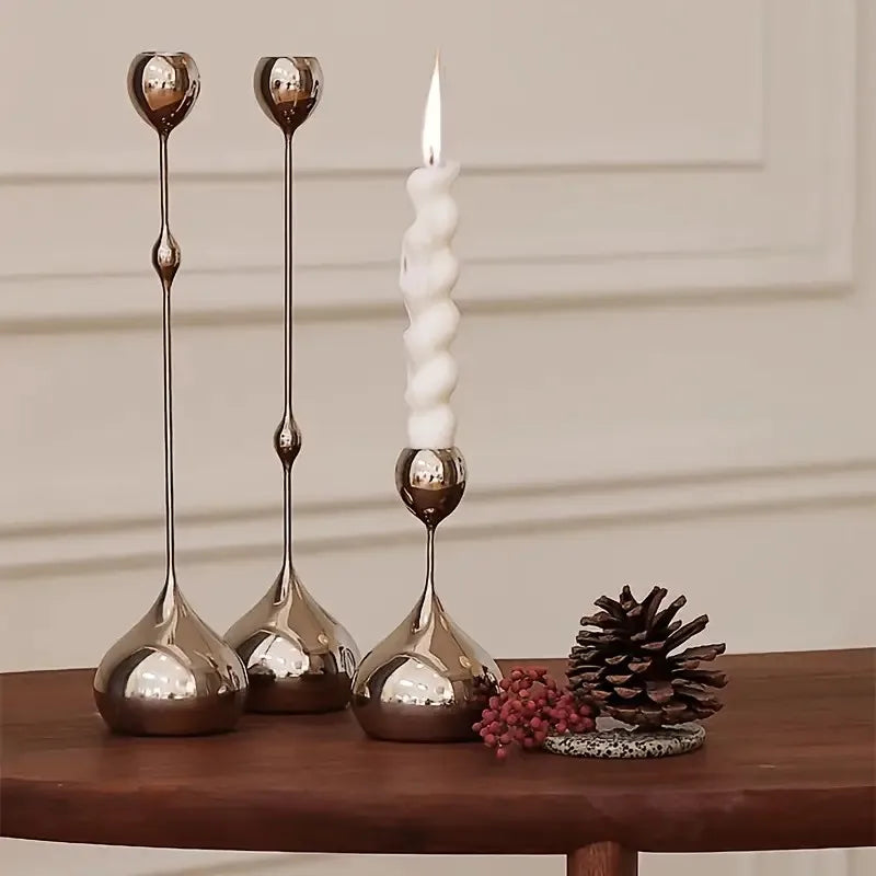 A set of three metallic waterdrop design candle holders with white candles and a pinecone on a wooden surface.