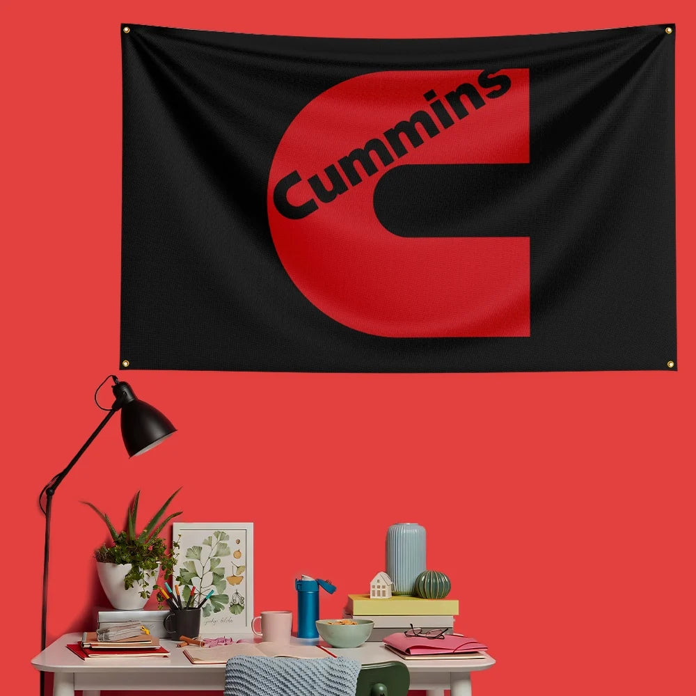 A black flag with the red letter C and the word Cummins printed on it, displayed on a red wall.