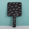 A black handheld vanity mirror with an ergonomic handle featuring a pattern of stylized eyelashes.