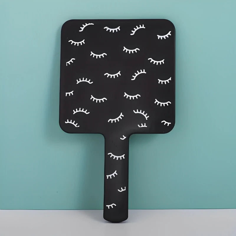 A black handheld vanity mirror with an ergonomic handle featuring a pattern of stylized eyelashes.