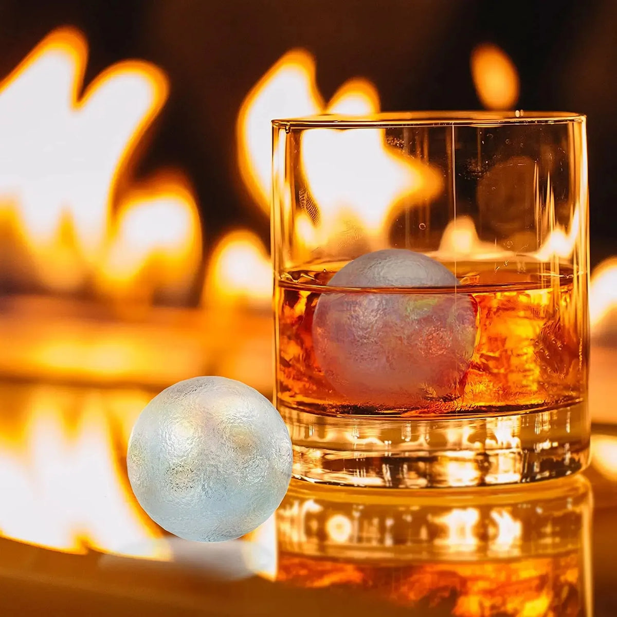 A glass of amber liquid with a silicone sphere and diamond ice mold resting on a glowing fire background.