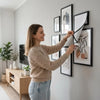 A woman is displaying a set of five black photo frames with white and natural-toned artwork on a white wall.