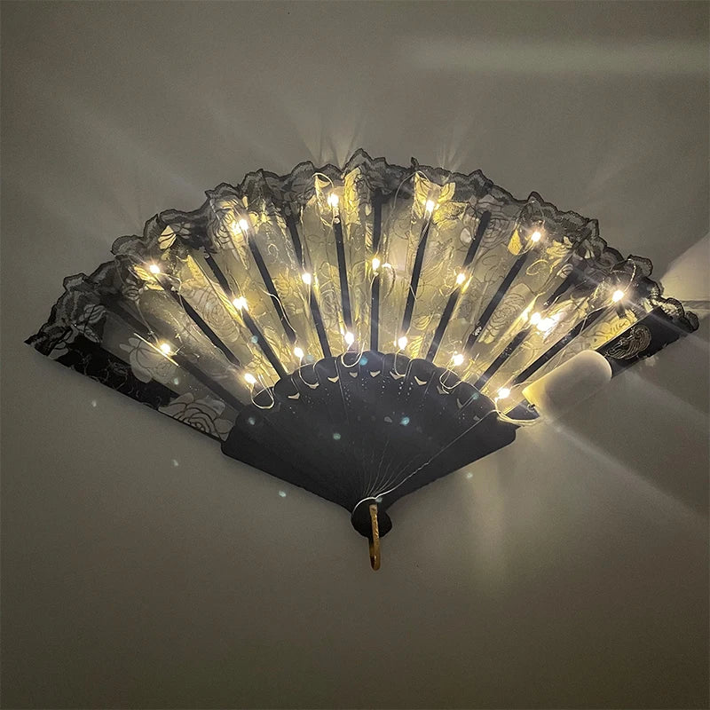 A black and gold LED dancing hand fan is illuminated, casting light rays against a neutral background.