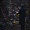 A person in a dark coat stands in a garden path, holding a decorative wind spinner made of dark feathers.