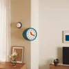 A modern resin wall clock with a blue face and orange hands is mounted on a beige wall.