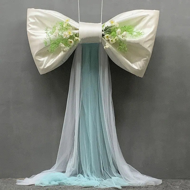 A white satin bow with green floral accents and a long, sheer light blue tulle drape for event decor.