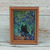 A framed painting featuring a black cat amidst blue and green flowers on a light wooden background.