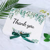 White gift box decorated with green eucalyptus leaves and a dark green ribbon tie.