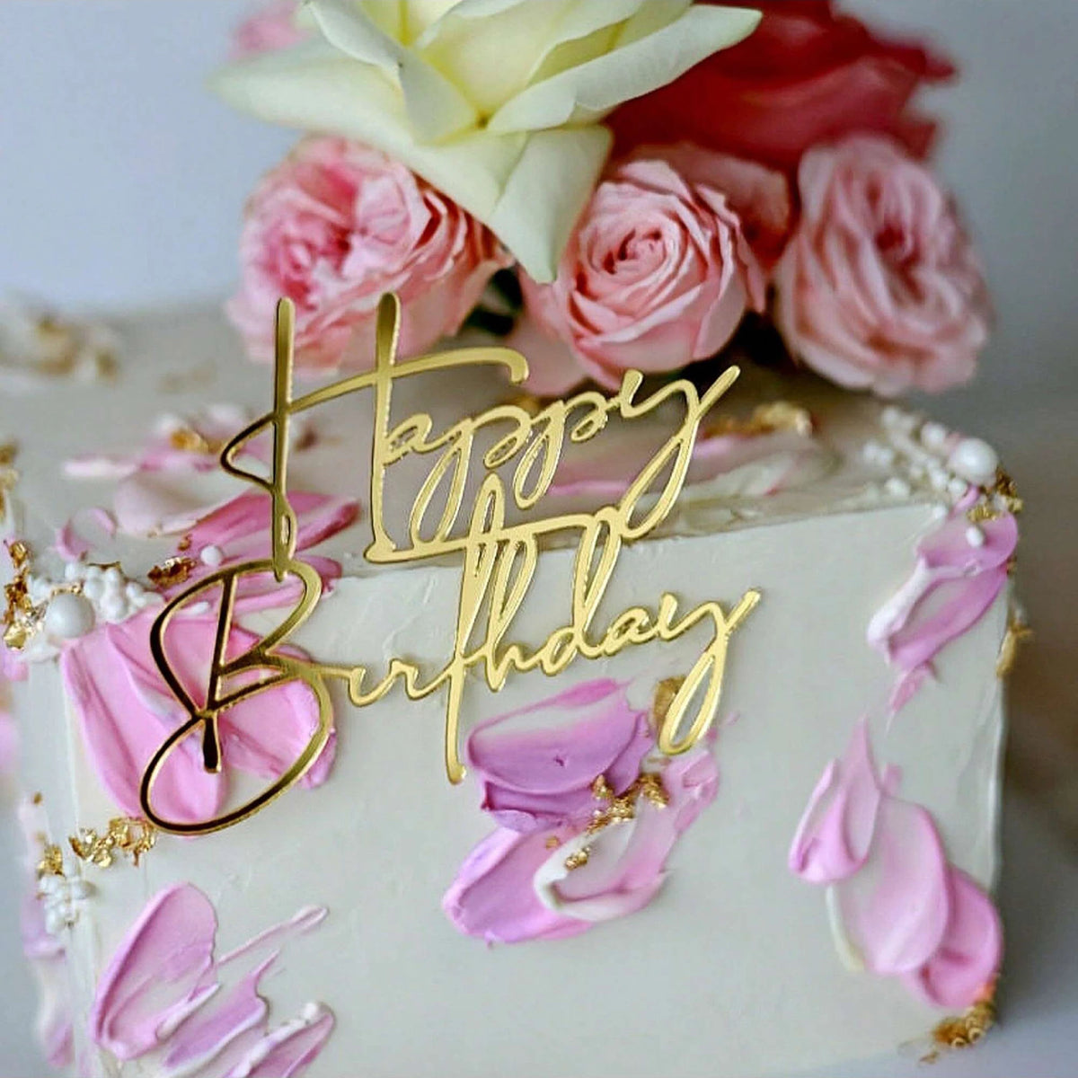 A white cake decorated with pink and gold floral accents and a gold acrylic happy birthday topper.