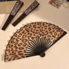 A leopard print folding hand fan with a black frame is displayed on a light surface.