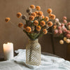 A vase filled with dried orange and brown chrysanthemum pompon flowers next to a lit candle on a white linen cloth.