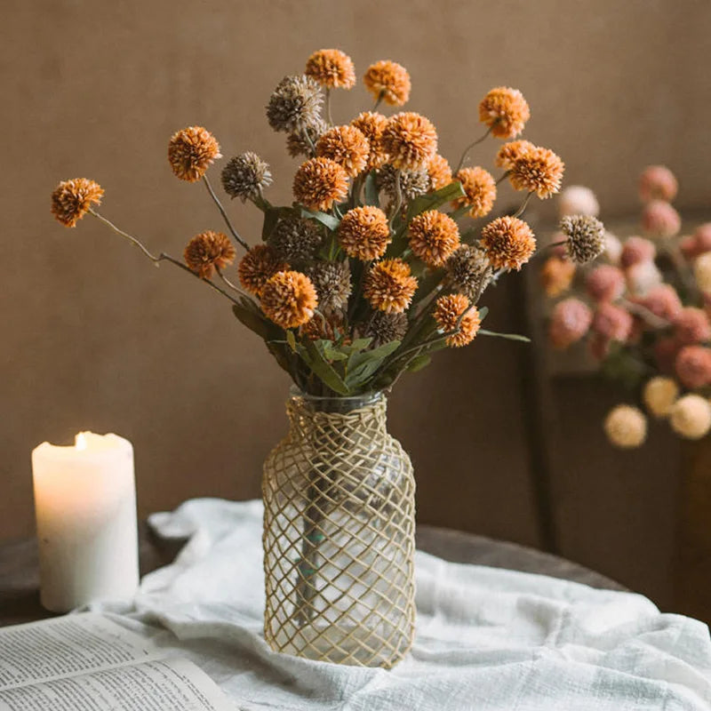 A vase filled with dried orange and brown chrysanthemum pompon flowers next to a lit candle on a white linen cloth.