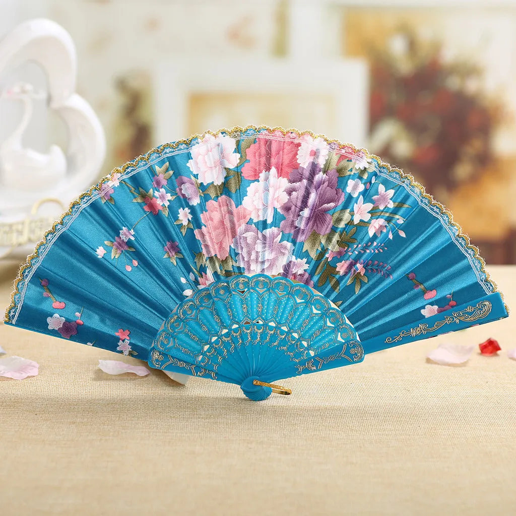 A teal hand fan decorated with pink and purple floral patterns, resting on a light beige surface.