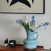 A light blue ceramic vase holds white and blue wildflowers on a wooden tray.