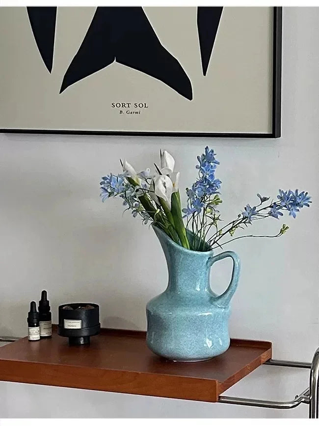 A light blue ceramic vase holds white and blue wildflowers on a wooden tray.