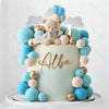 A round light blue cake decorated with blue and beige foam balls and a teddy bear topper.