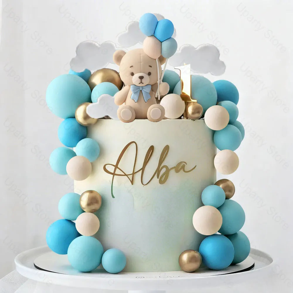 A round light blue cake decorated with blue and beige foam balls and a teddy bear topper.