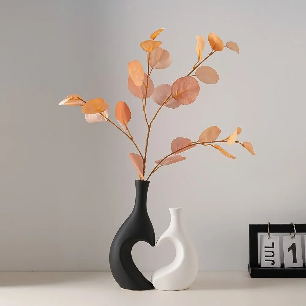 A modern heart-shaped ceramic vase holds delicate dried leaves in soft peach and beige tones.