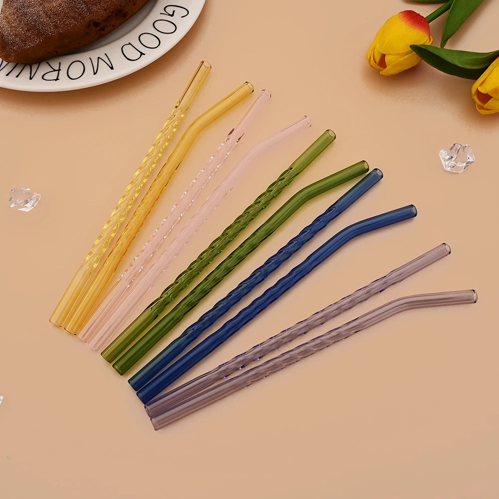A collection of various colored reusable borosilicate glass straws with cleaning brushes arranged on a light background.