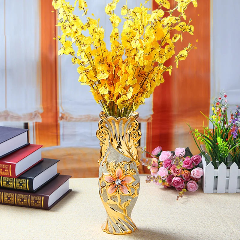 A gold-plated ceramic tabletop vase filled with bright yellow flowers on a light-colored table.