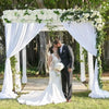 A bride and groom stand under a white floral arch panel decorated with greenery in an outdoor setting.