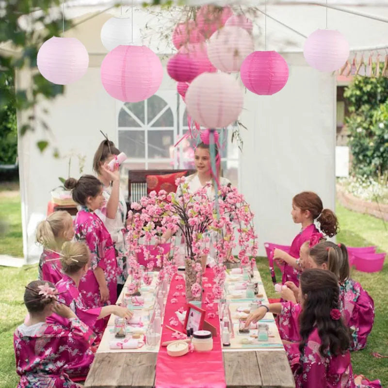 Children in pink floral dresses participate in a celebration under white draping with pink paper lanterns.